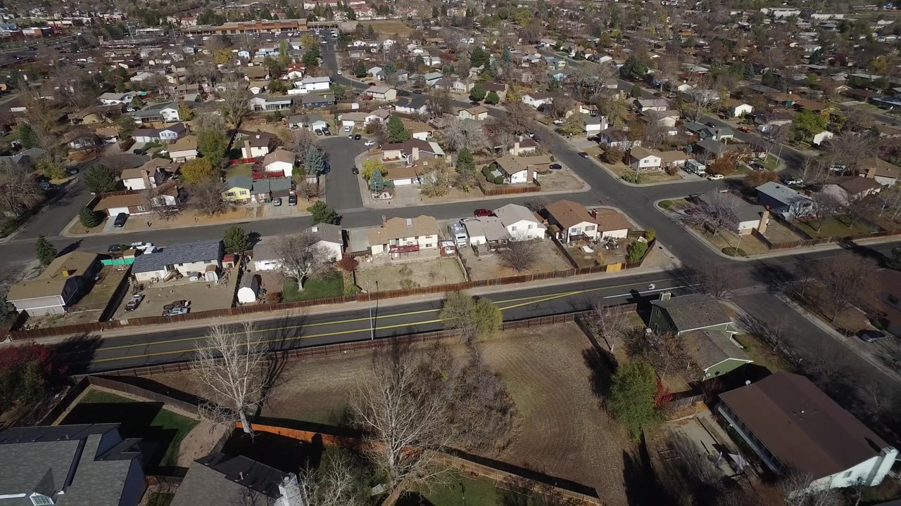 Oblique aerial frame over Hidden Lake, Colorado houses, fenced yards, a main road, leafless trees, and no visible people.