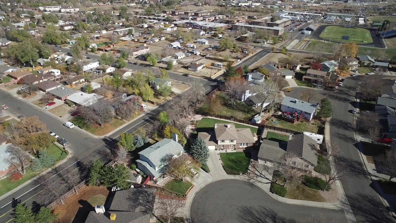 Aerial frame of Hidden Lake, Colorado homes around a cul-de-sac, with rooftops, yards, leafless trees, a broad road, and the school field farther back, with no visible people.