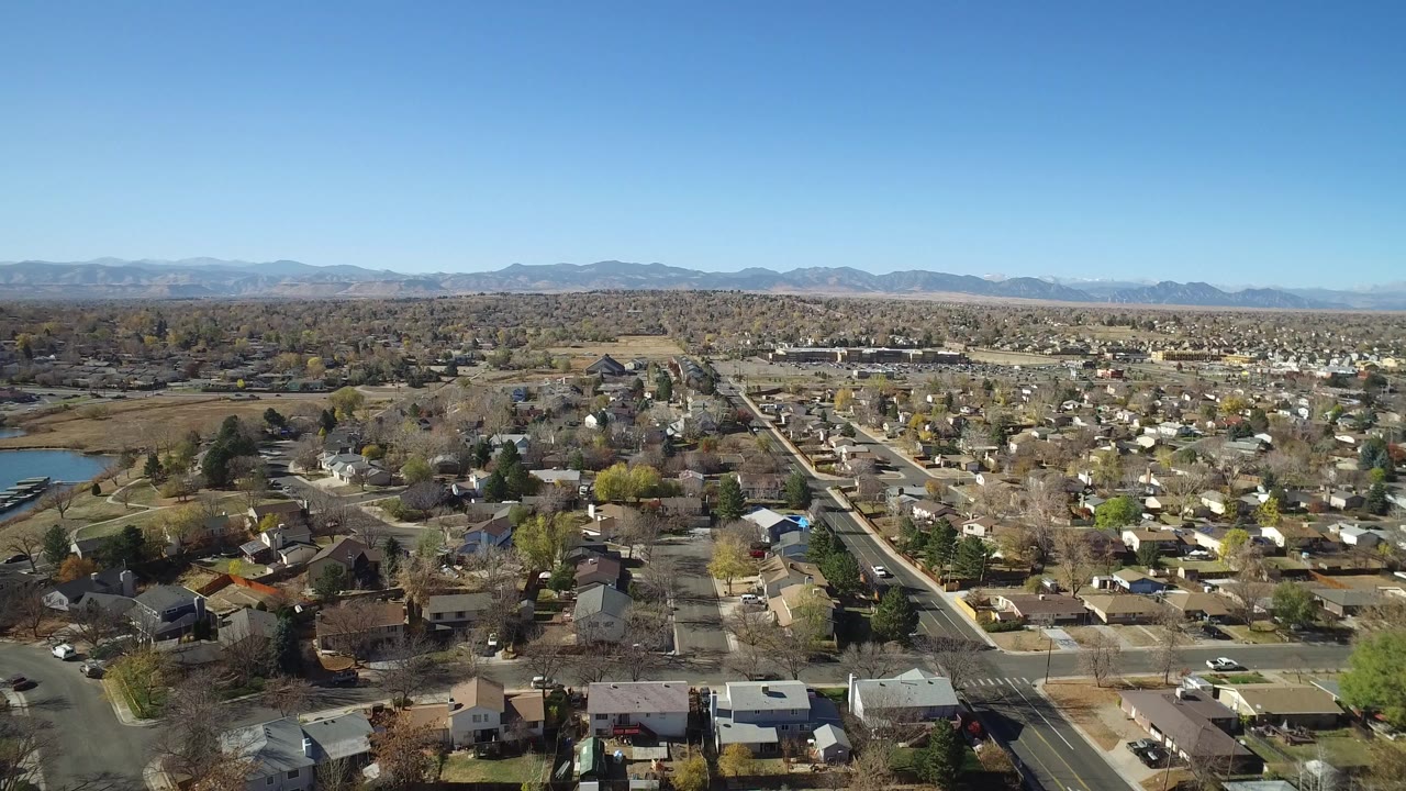 Aerial view of suburban homes, streets, trees, water, and distant mountains under a clear sky.