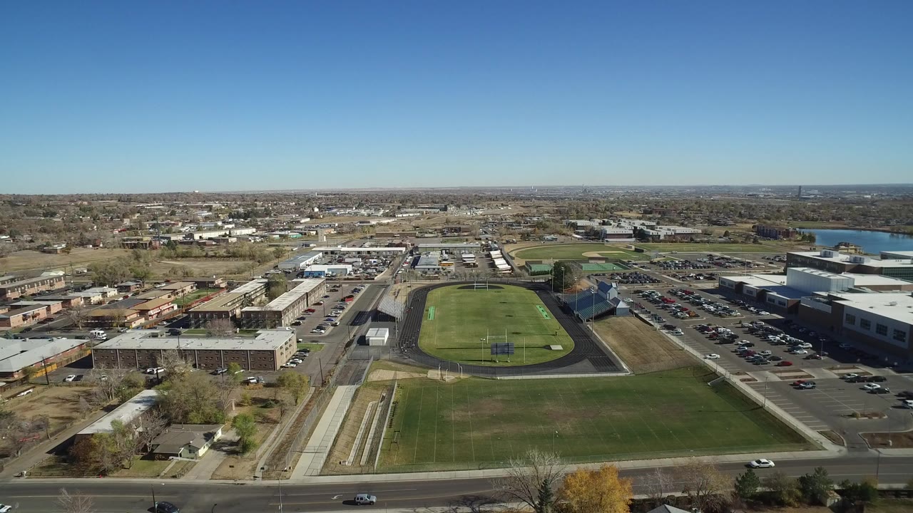 Wide drone view over Westminster, Colorado showing a green athletic field inside a running track, nearby roads, parking lots, low buildings, distant neighborhoods, and no visible people.