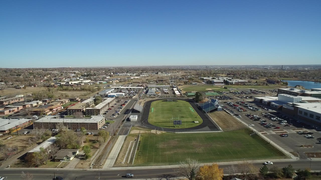 Drone frame over Westminster, Colorado with the running track and field centered, a large parking lot to the right, low retail-style buildings to the left, distant housing, and no visible people.