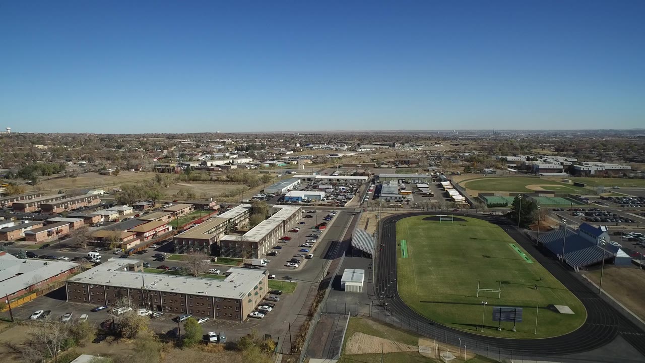 Aerial frame in Westminster, Colorado showing the green field slightly right of center, a long building in the foreground, parked cars, and no visible people.