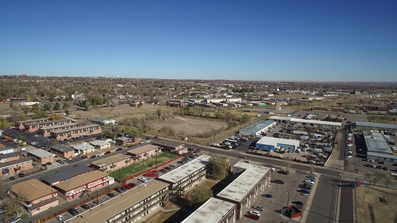 Aerial view over apartment buildings, streets, parked cars, and distant houses under a blue sky.