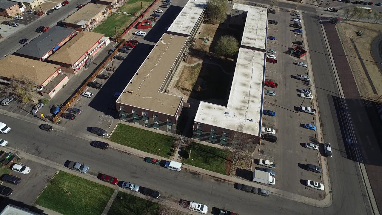 High aerial view in Westminster, Colorado over flat-roof buildings, parked cars, paved streets, small grass patches, and no visible people at the opening moment.