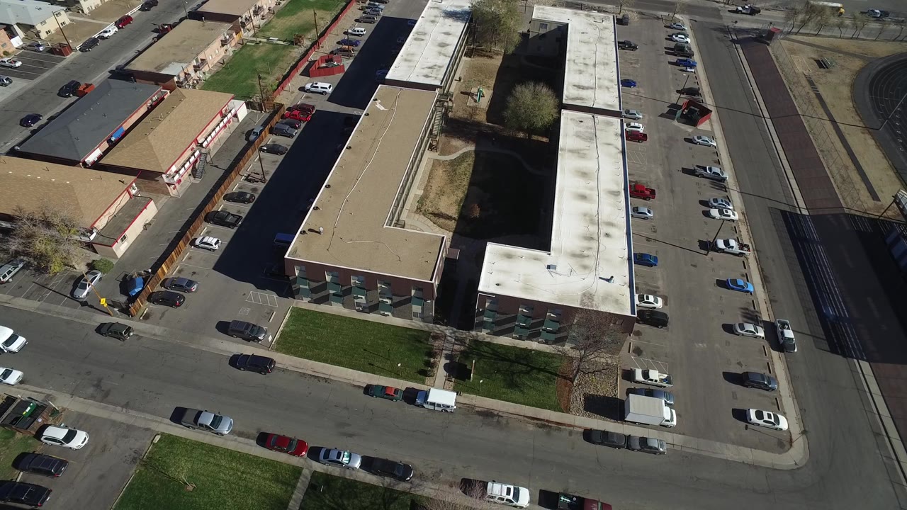 Aerial view of buildings, parked cars, road lanes, and a rail or road corridor.