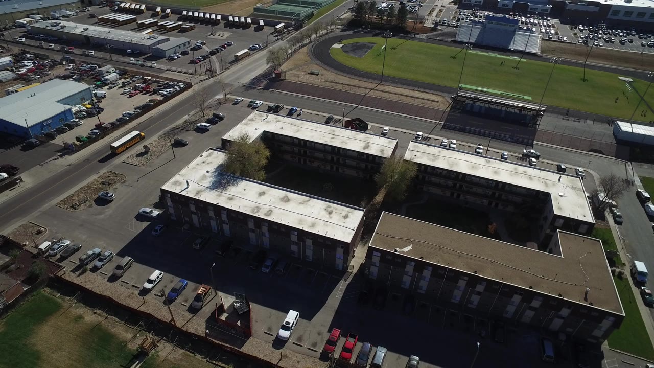High aerial frame over Westminster, Colorado showing a large condominium building, parked cars, surrounding roads, lawns, and a track field in the background with no visible people.