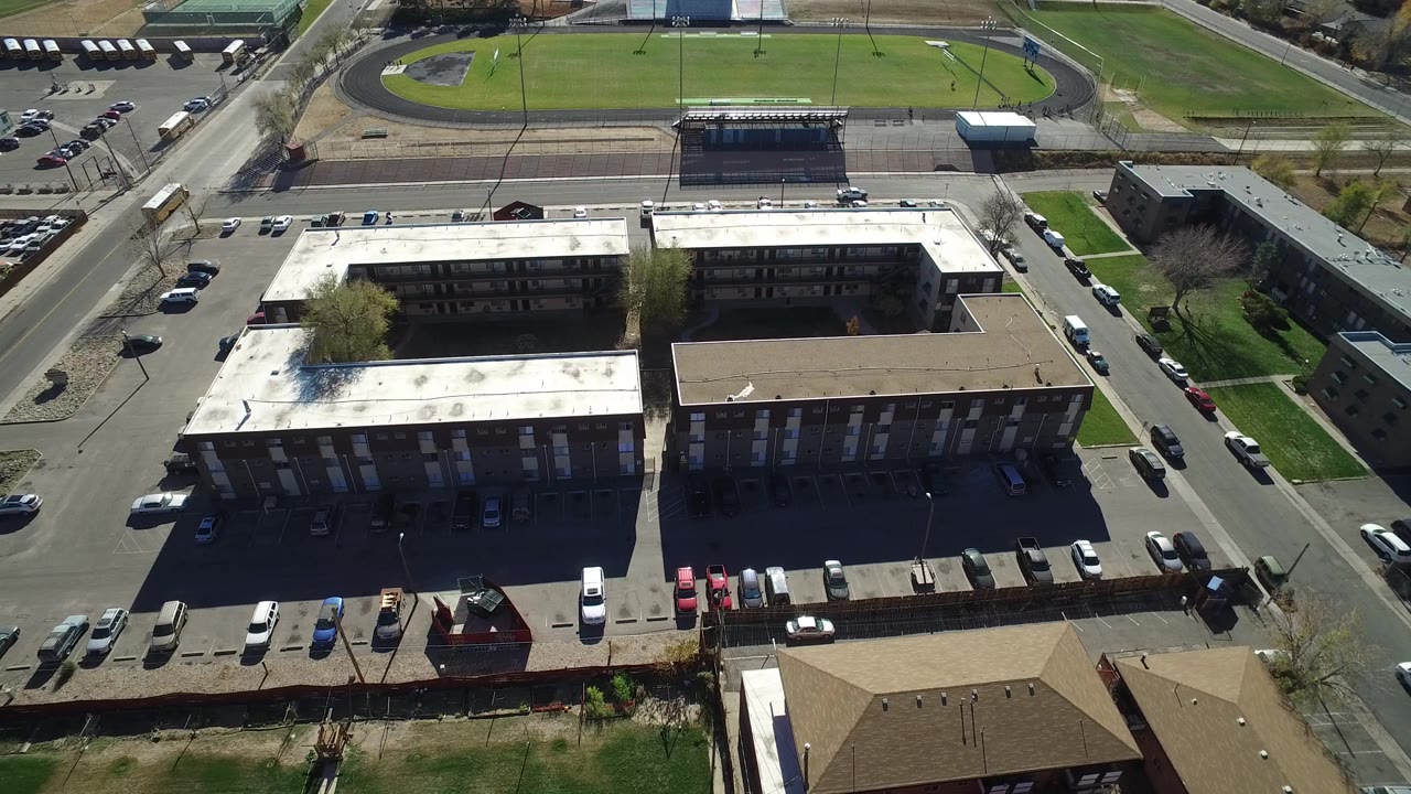 High aerial frame over Westminster, Colorado with the bright white roof on the left building, a tan roof on the right building, parked cars, streets, and a track beyond, with no visible people.