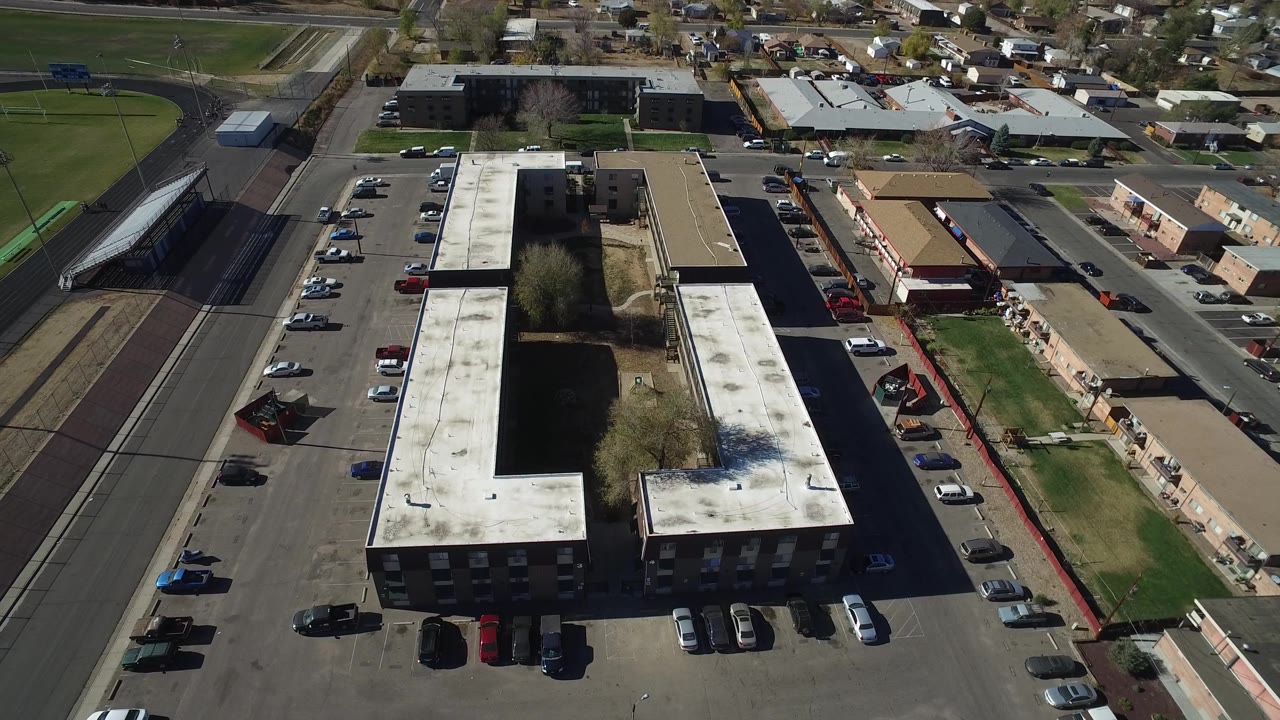 Aerial view in Westminster, Colorado with the white roof of a large building centered, parking spaces and cars around it, a road to the right, and homes beyond, with no visible people.