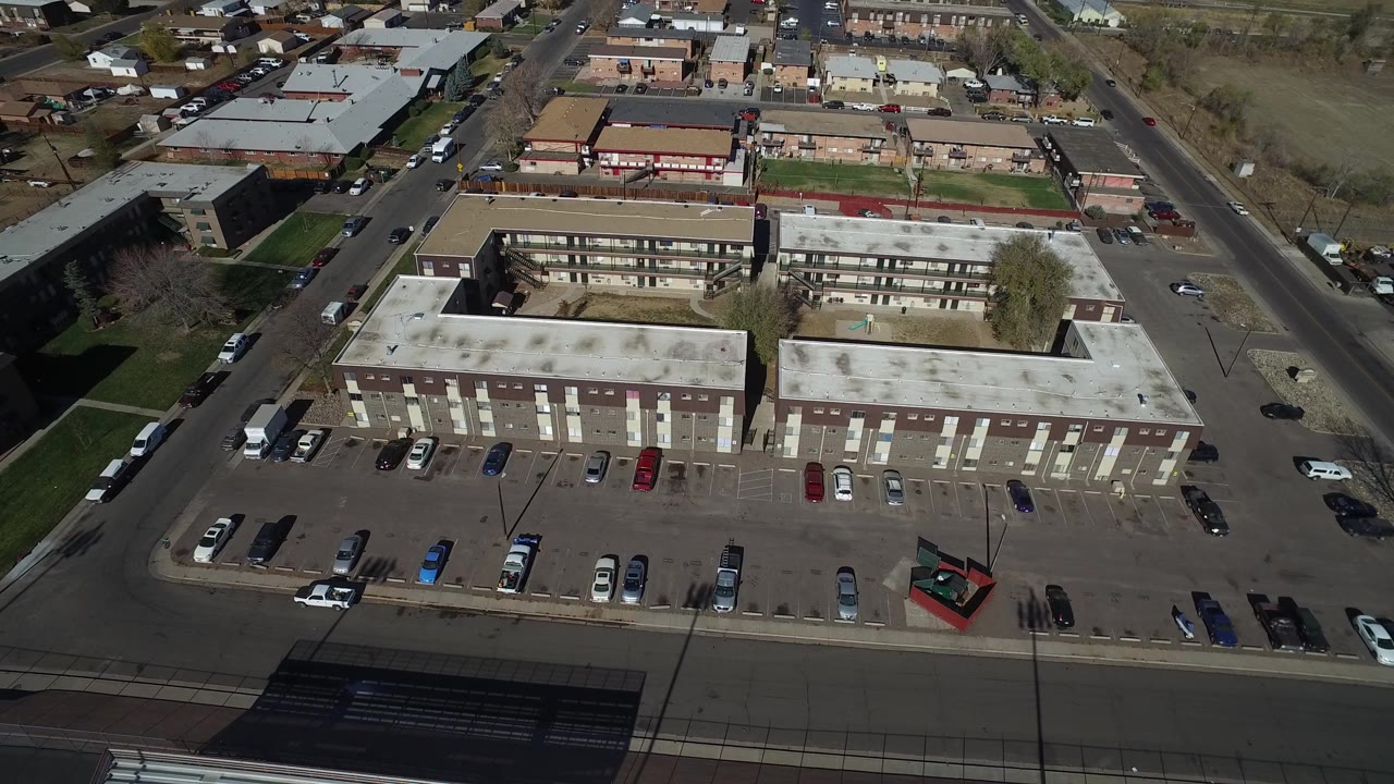 High aerial view over Westminster, Colorado low-rise buildings with flat roofs, rows of parked cars, surrounding streets, and no visible people at the start of the clip.