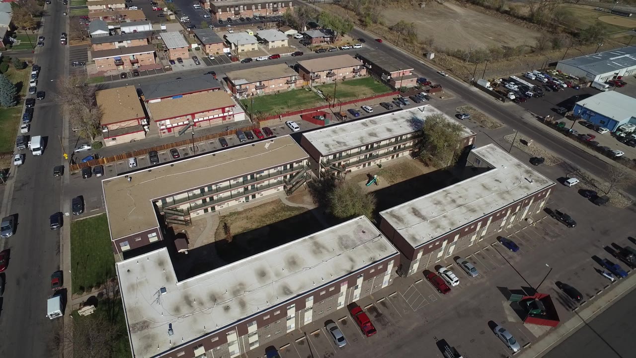 Aerial frame in Westminster, Colorado with the main flat-roof building centered lower in frame, small commercial buildings across the road, parking rows, and no visible people.