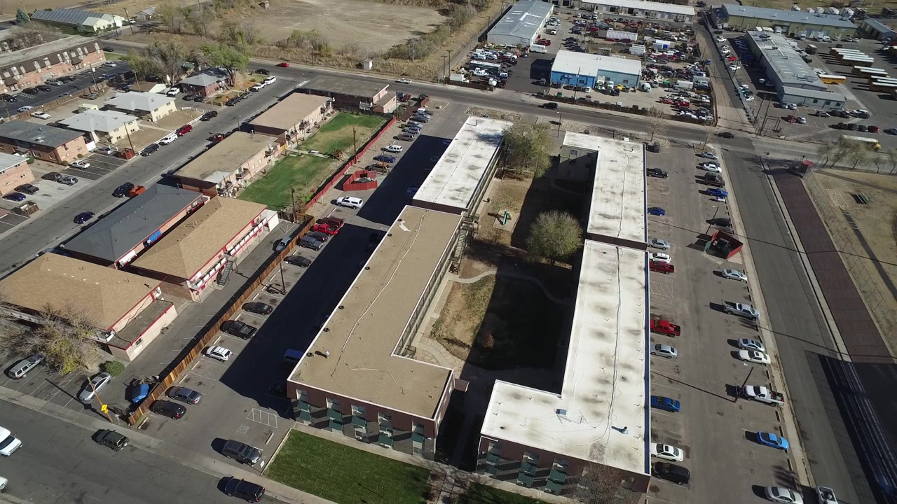 Aerial view in Westminster, Colorado, with the tan building block and white-roofed structure filling the center, roads and parked vehicles around them, and no visible people.