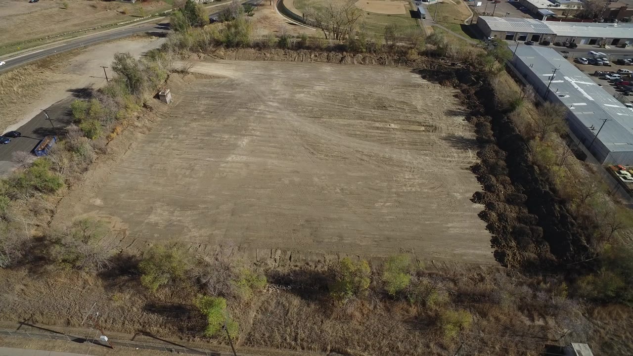 High drone view in Westminster, Colorado, with the brown cleared field centered, brush along the near edge, buildings on the right side, and no visible people.