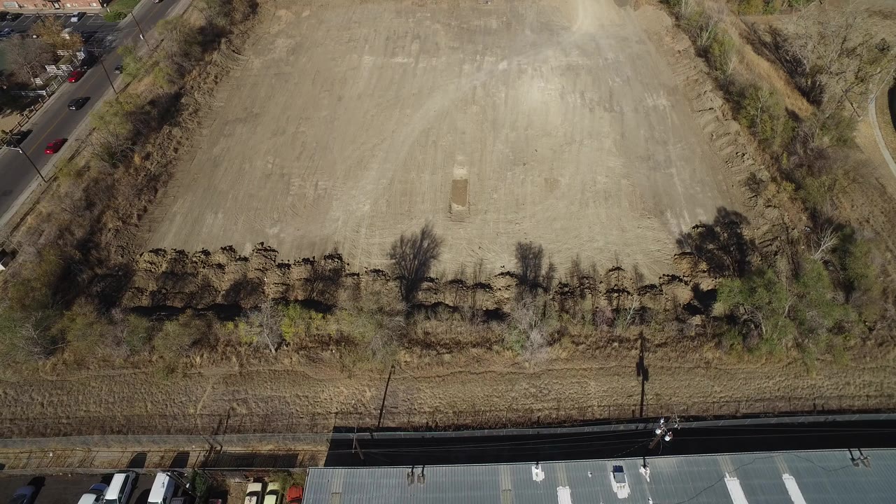 Overhead aerial frame in Westminster, Colorado, showing the tan field centered between roadside edges, leafless trees, scrub vegetation, and no visible people.