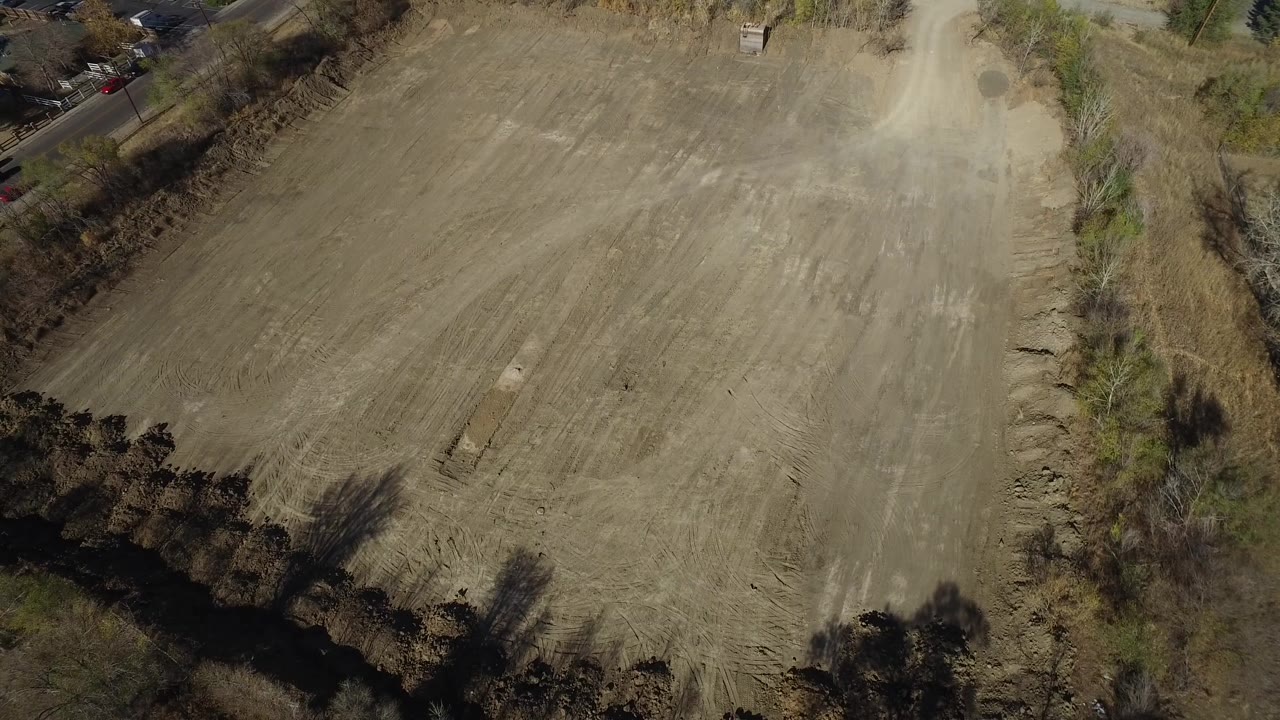 High aerial view over a plowed field in Westminster, Colorado, with bare trees along the lower edge, a road at the upper left, and no visible people.