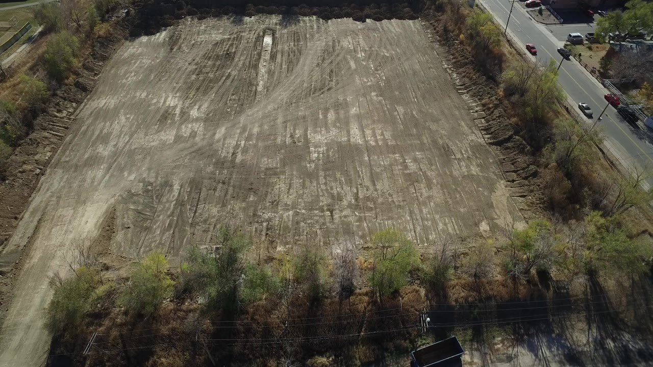 High aerial view over a large tan dirt plot in Westminster, Colorado, with trees along the edges, a paved road at right, and no visible people.