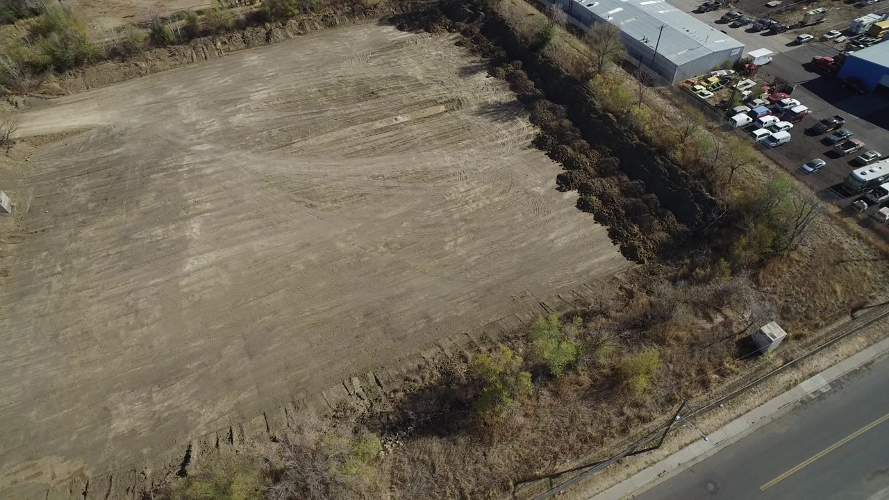 A high aerial view in Westminster, Colorado shows a large cleared dirt field bordered by brush, a road at the lower edge, warehouse roofs at the upper right, and no visible people.