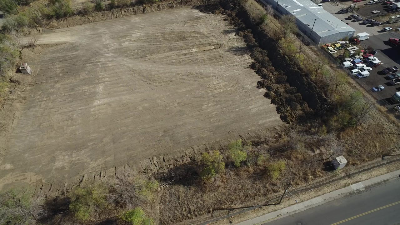 Drone view of an empty field with brush, roadside pavement, and parked vehicles by a warehouse.
