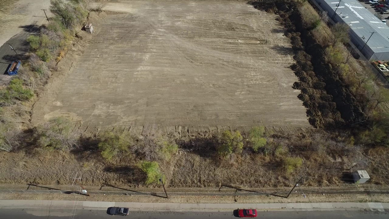 Overhead frame in Westminster, Colorado showing a dry open field, roadside trees, a paved street with two cars, and no clearly identifiable people.