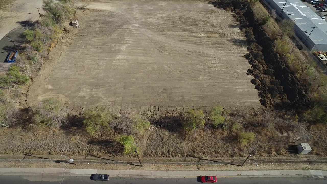 High drone view in Westminster, Colorado with the field centered, a tree line along the road, cars moving along the lower edge, and no clearly identifiable people.