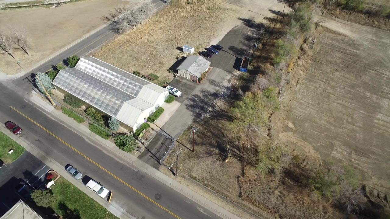 Aerial frame in Westminster, Colorado with a red car on the road, the greenhouse-like building at left, open tan field at right, and no visible people.