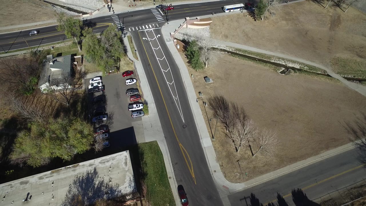 Aerial frame in Westminster, Colorado showing a broad road, a side street, a small row of parked vehicles, dry grass, sidewalks, trees, and no visible people.