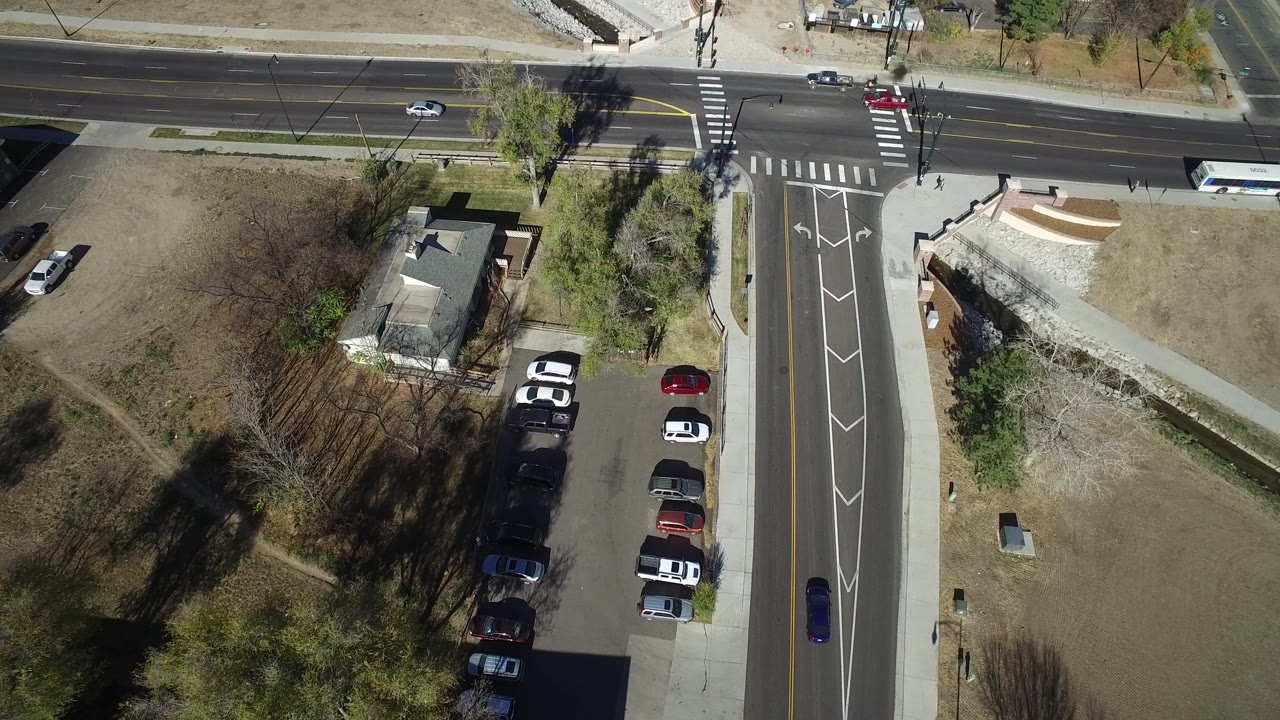 Overhead frame from Westminster, Colorado showing a road intersection with zebra crossings, a small building, parked cars, dry grass, trees, and tiny pedestrians near the upper crosswalk.