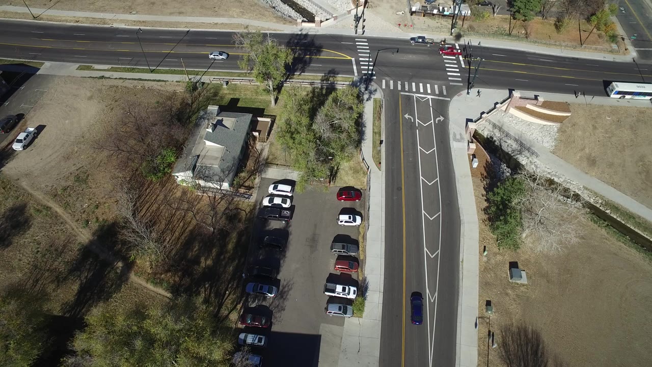 Aerial view in Westminster, Colorado with a bus moving along the main road, cars near the crosswalks, a perpendicular lane with arrows, trees, and parked vehicles beside a small building.