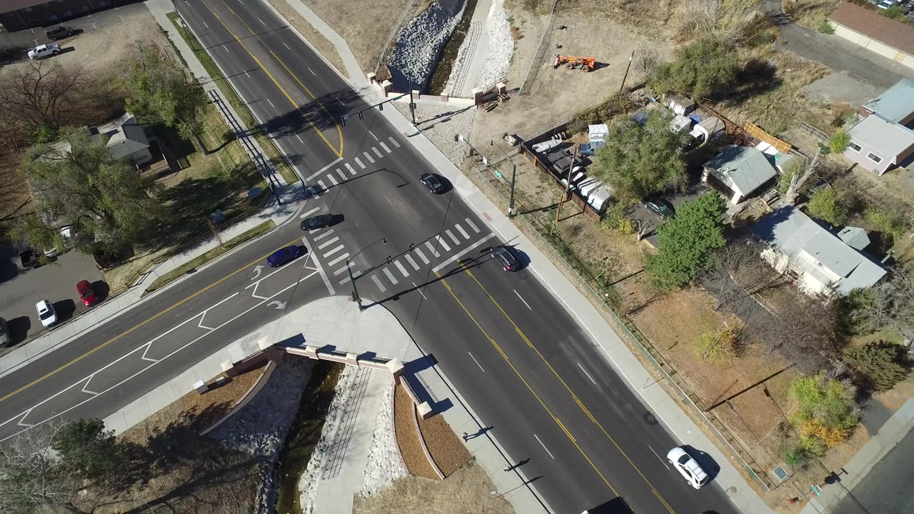 Aerial frame from Westminster, Colorado with vehicles spaced across the road, striped crosswalks, lane arrows, residential lots, trees, sidewalks, and no visible people.