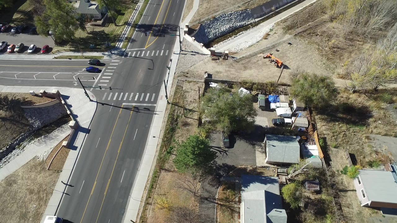 Aerial frame over Westminster, Colorado showing a paved intersection, marked crosswalks, sidewalks, trees, parked cars, and a drainage channel, with no visible people.