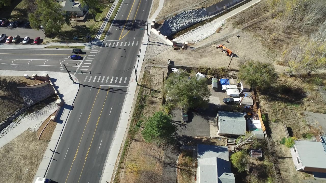 A high overhead view in Westminster, Colorado shows the road junction shifting across frame with crosswalk stripes, sidewalks, a rock-lined water channel, parked vehicles, and no visible people.