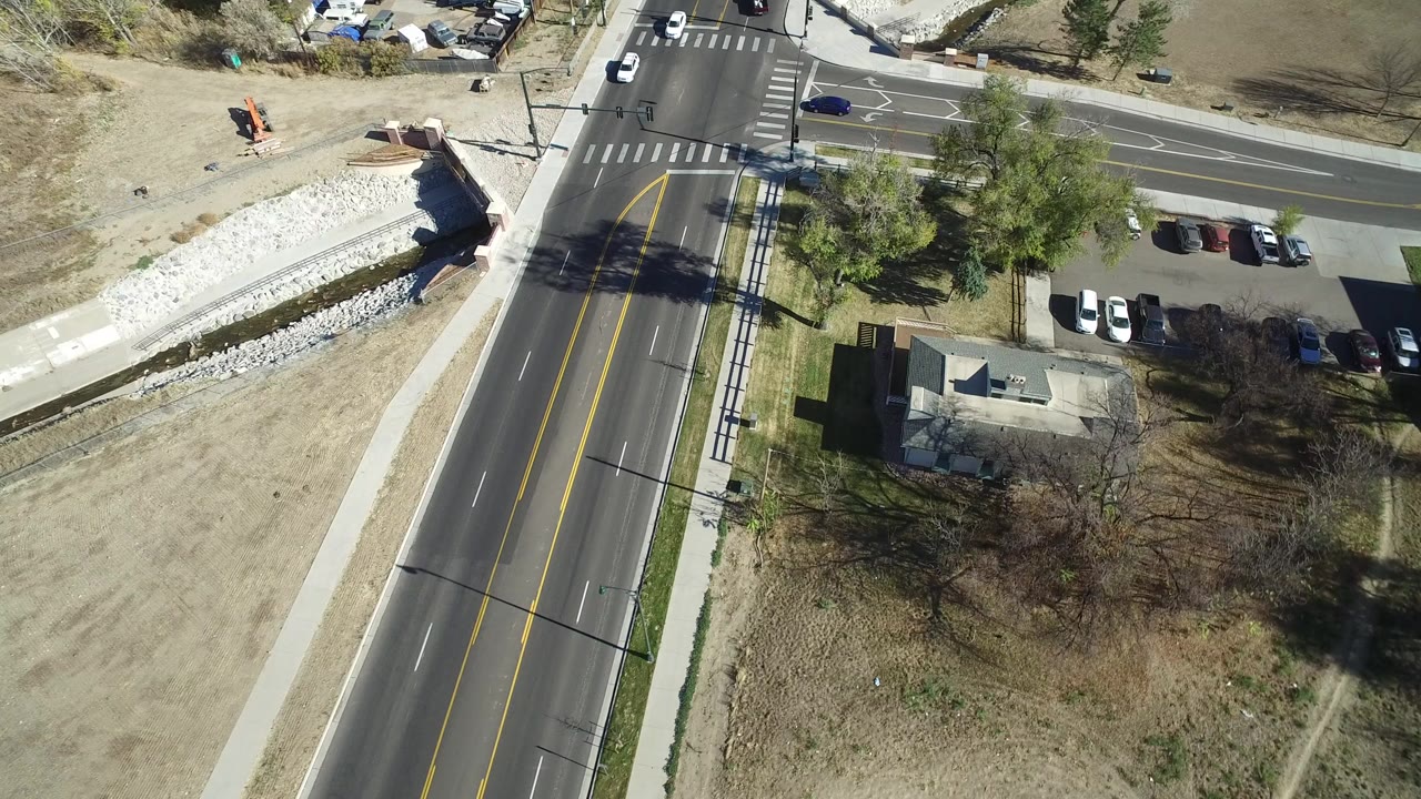 Overhead aerial frame in Westminster, Colorado showing a diagonal road, adjacent side street, parked cars, a small building, dry grass, trees, and no visible people.