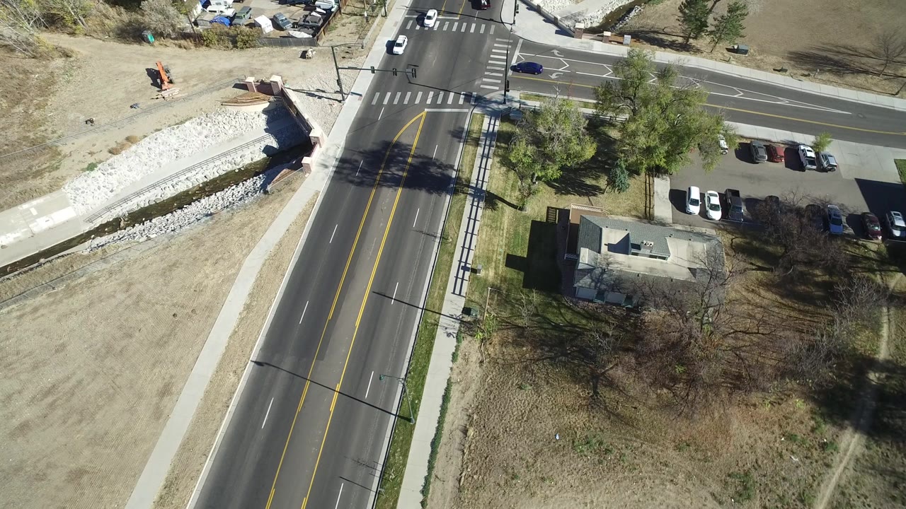 Westminster, Colorado aerial frame looking down on a road and side street junction with lane markings, sidewalks, parked cars, tree shadows, dry open ground, and no visible people.