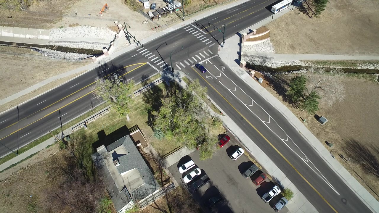 Aerial view of a road intersection beside grass, trees, parked cars, and a small building.