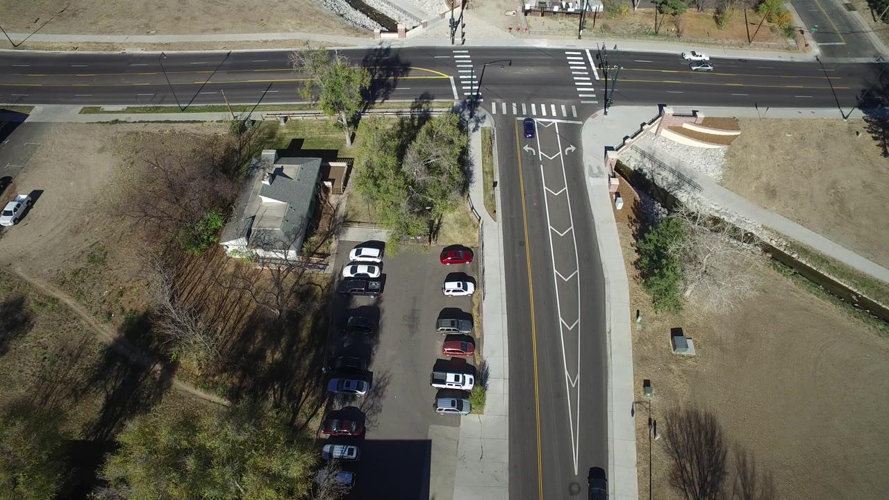 Overhead frame of a Westminster, Colorado road intersection with crosswalks, turn lanes, parked cars, a small building, dry grass, trees, and no visible people.