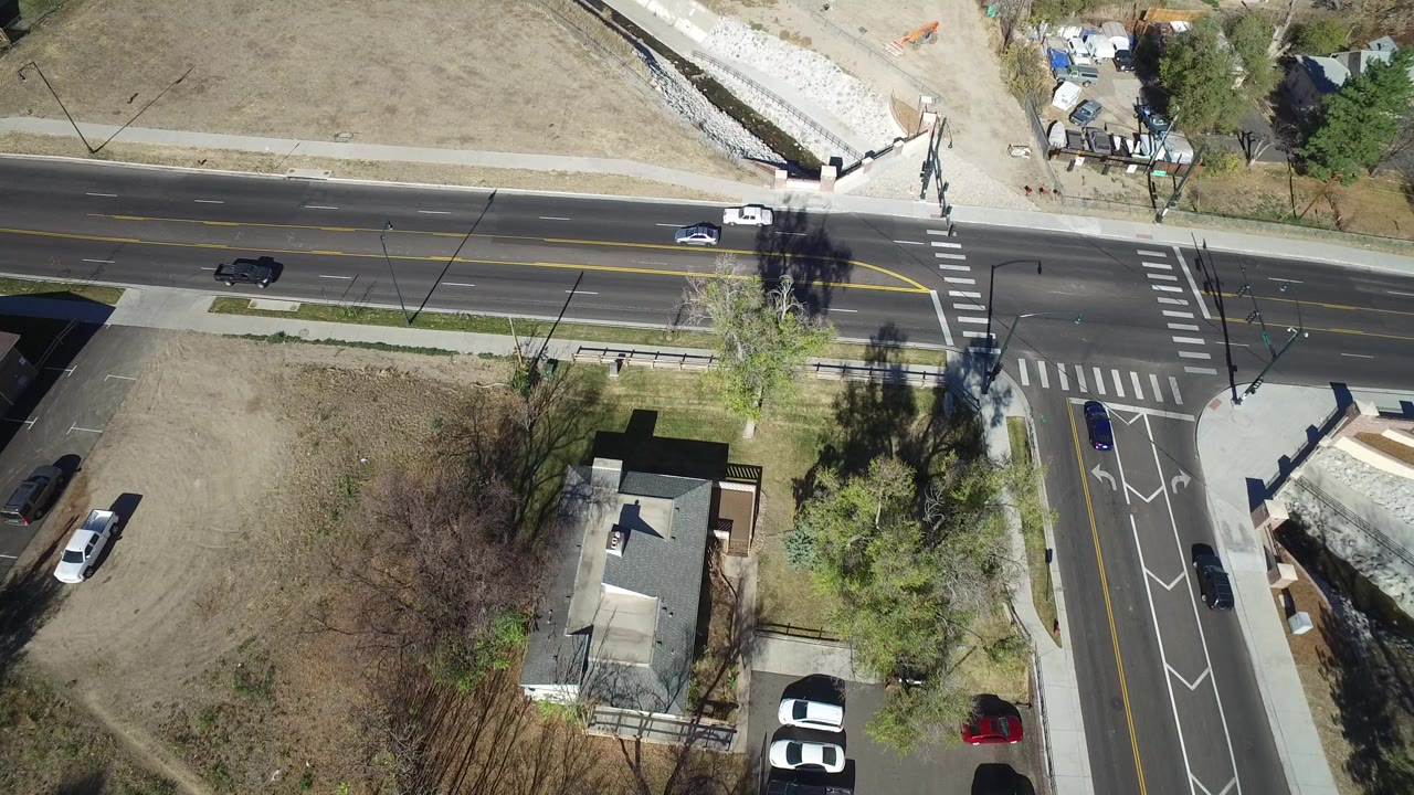 Overhead view in Westminster, Colorado showing a multi-lane road, marked crosswalks, a side street, open dry land, trees, nearby homes, several cars, and no visible people.