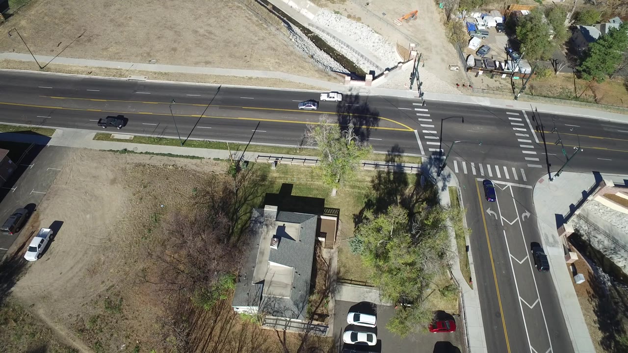 Aerial frame in Westminster, Colorado with cars on a multi-lane road, a crosswalk at a side street, dry open ground, trees, residential houses, and no visible people.