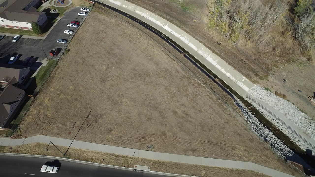 Top-down aerial frame in Westminster, Colorado showing open brown grassland bordered by homes, a road, a sidewalk, a drainage channel, parked cars, and no visible people.