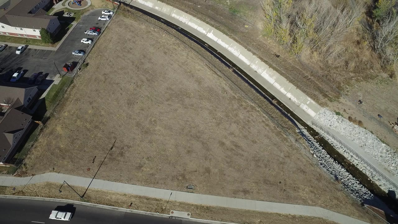 Aerial view in Westminster, Colorado with a white vehicle on a dark road, open grassland, sidewalks, residential buildings, a concrete channel, and no visible people.