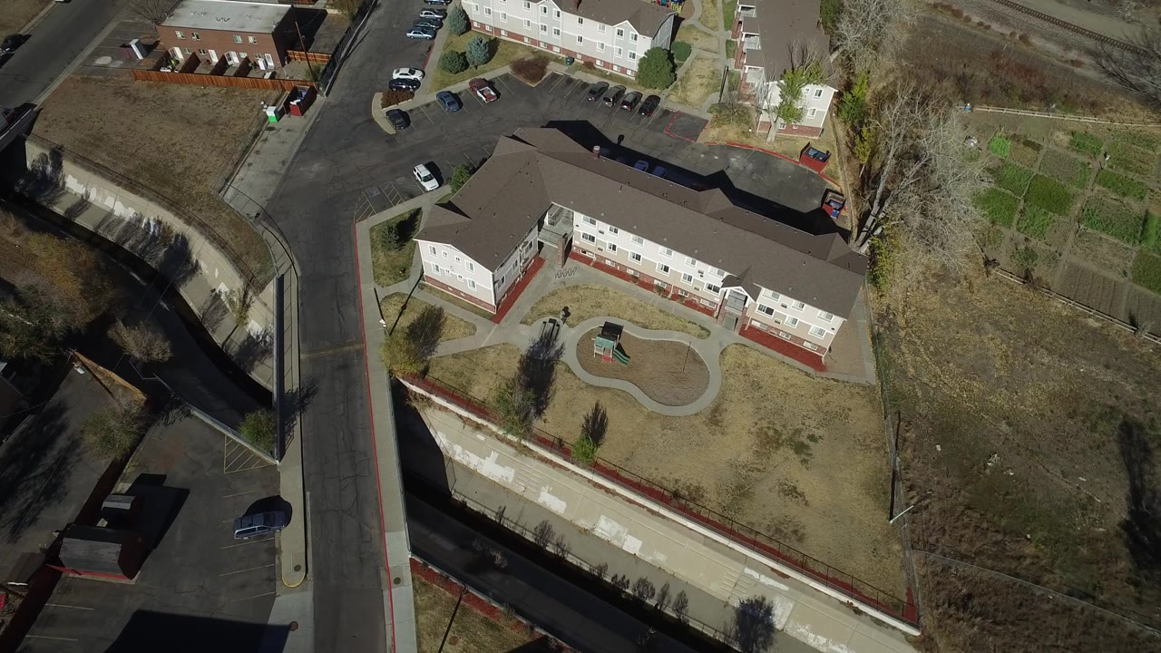 Aerial view in Westminster, Colorado showing a long residential building, adjacent road, parked cars, open dry ground, trees, and no visible people.