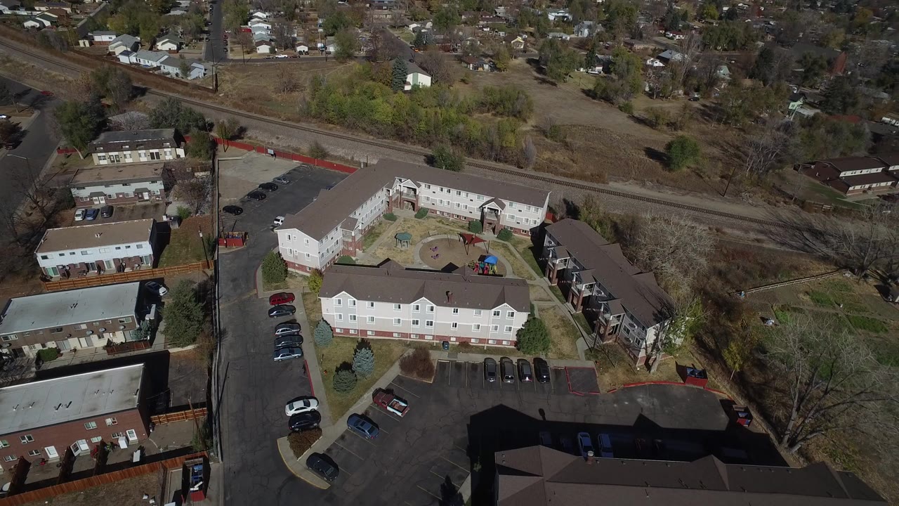 High aerial frame of Westminster, Colorado residential buildings, a central courtyard, parked cars, a road and rail line across the upper frame, and no visible people.