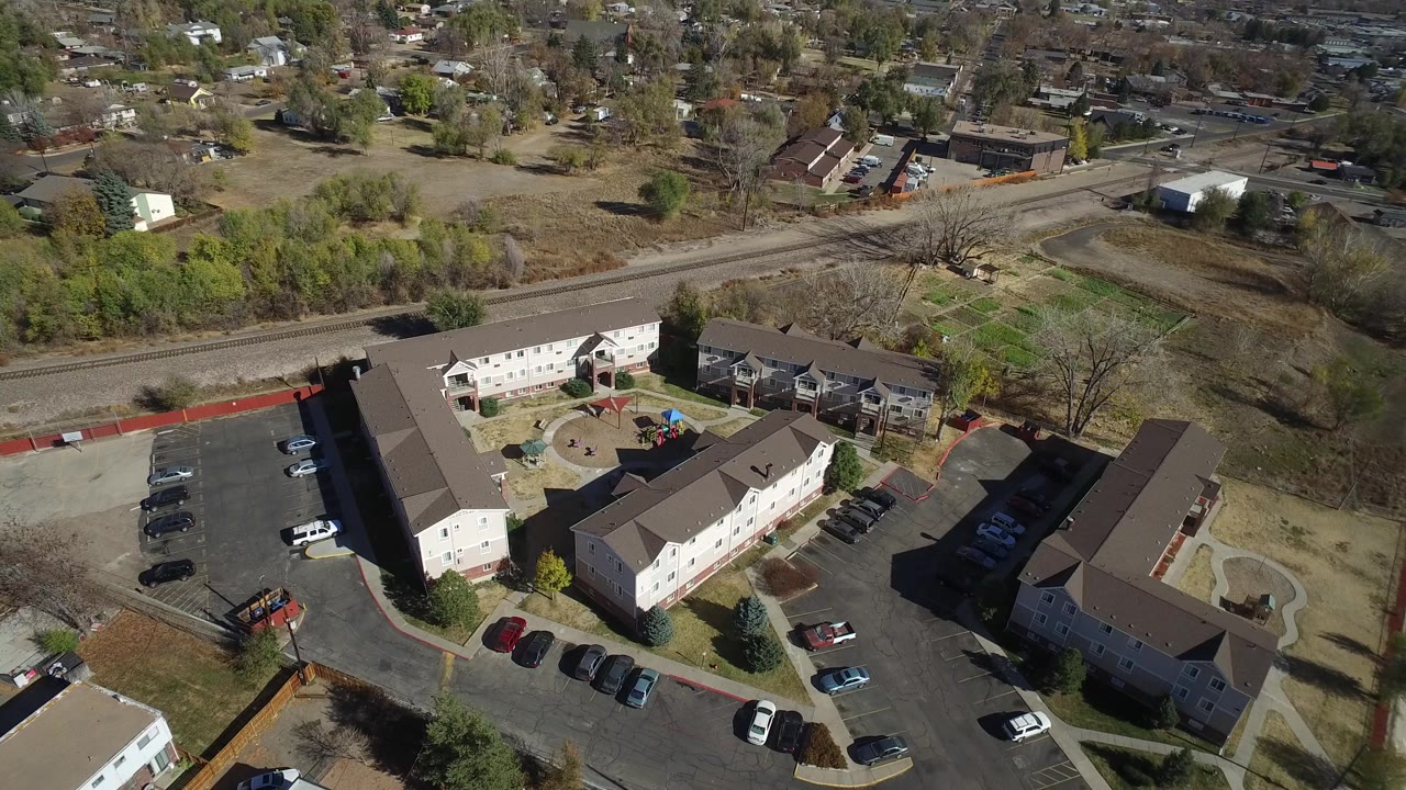 Aerial frame over Westminster, Colorado apartments with a small play area or courtyard near the center, parked vehicles below, rail tracks behind, and no visible people.