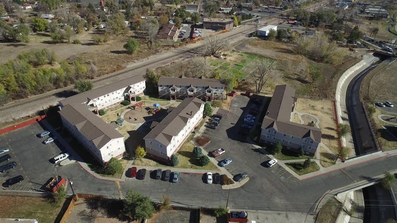 Aerial frame in Westminster, Colorado with the building complex centered lower in view, rows of parked cars, a road intersection, open ground, trees, and no visible people.