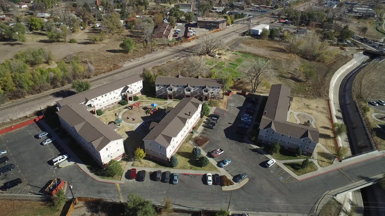 Elevated drone-style view in Westminster, Colorado showing a courtyard between multi-story buildings, parked vehicles, a curving road, sparse trees, dry grass, and no visible people.