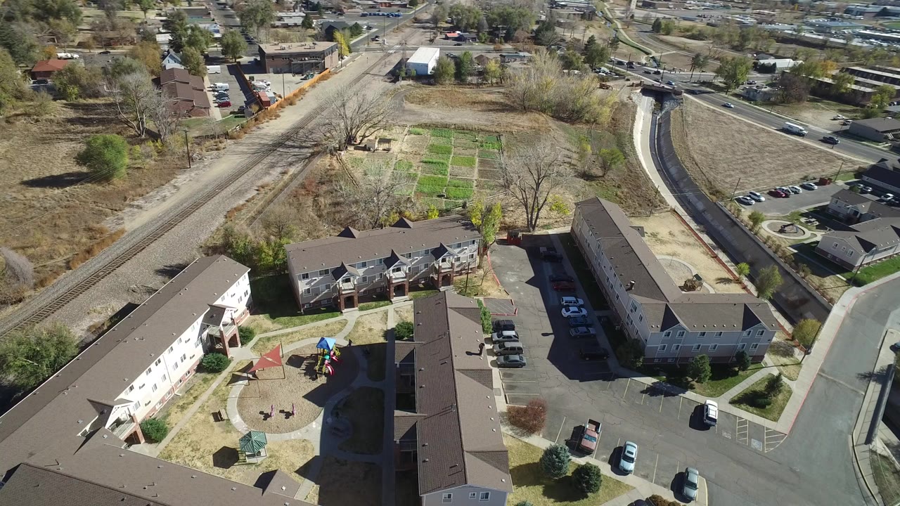 Aerial frame over Westminster, Colorado showing apartment buildings around a courtyard, parked cars, nearby roads, railroad tracks, dry open lots, and no visible people.