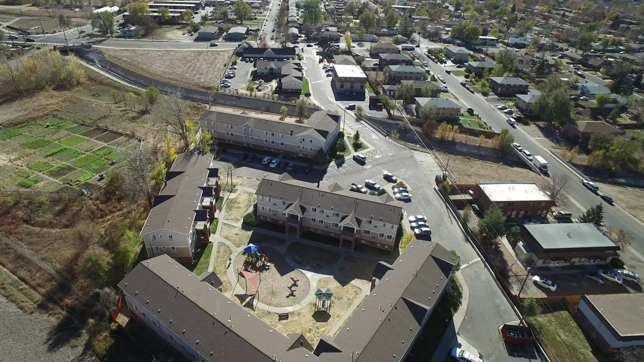 Aerial view in Westminster, Colorado looking down on multi-story homes, a curving street, parked cars, green garden plots, bare trees, and no visible people.