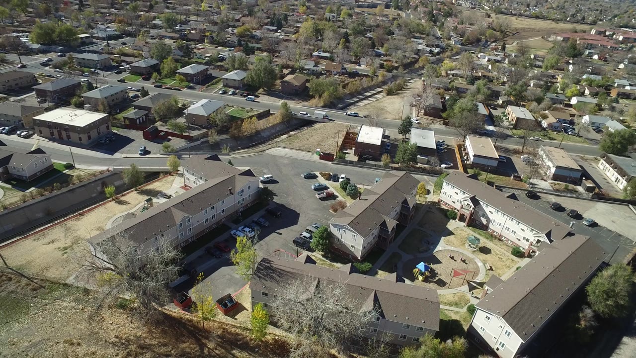 Aerial view in Westminster, Colorado with apartment roofs crossing the lower frame, parked cars near small lots, houses in the background, and no visible people.