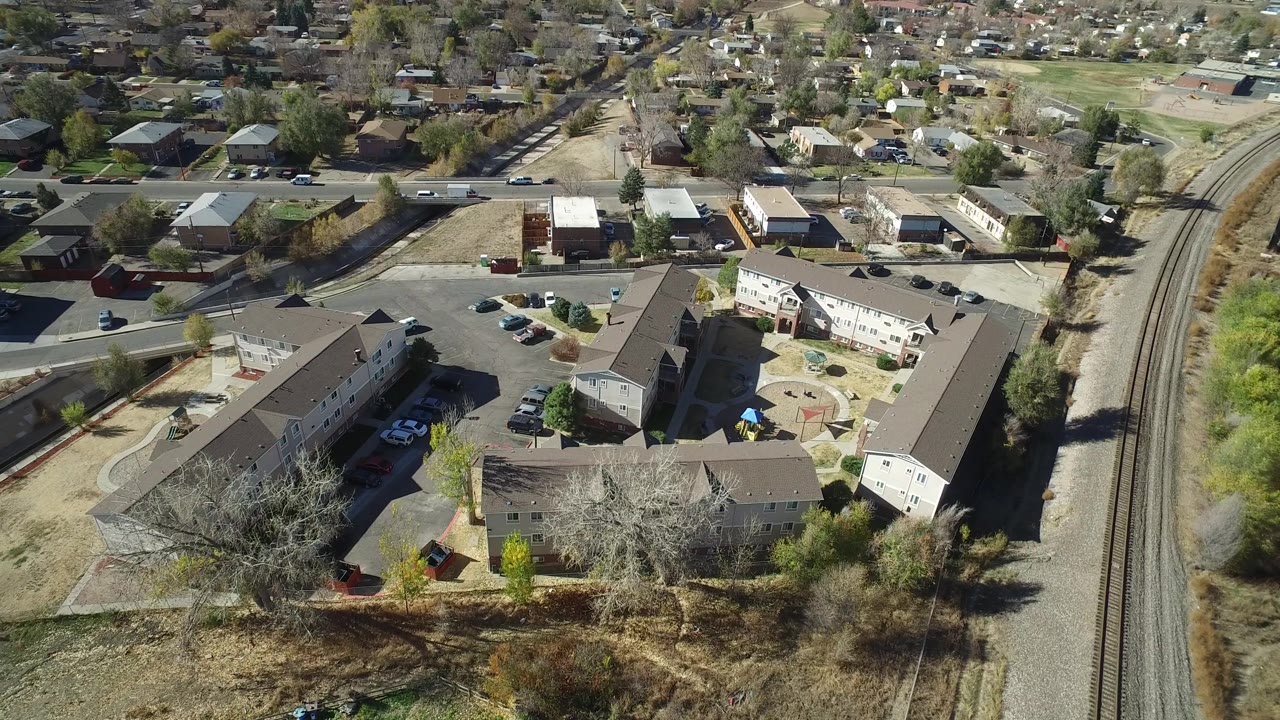 At 0:01, the Westminster aerial view keeps the rail tracks on the right with the building complex, parking areas, houses, and no visible people in bright daylight.