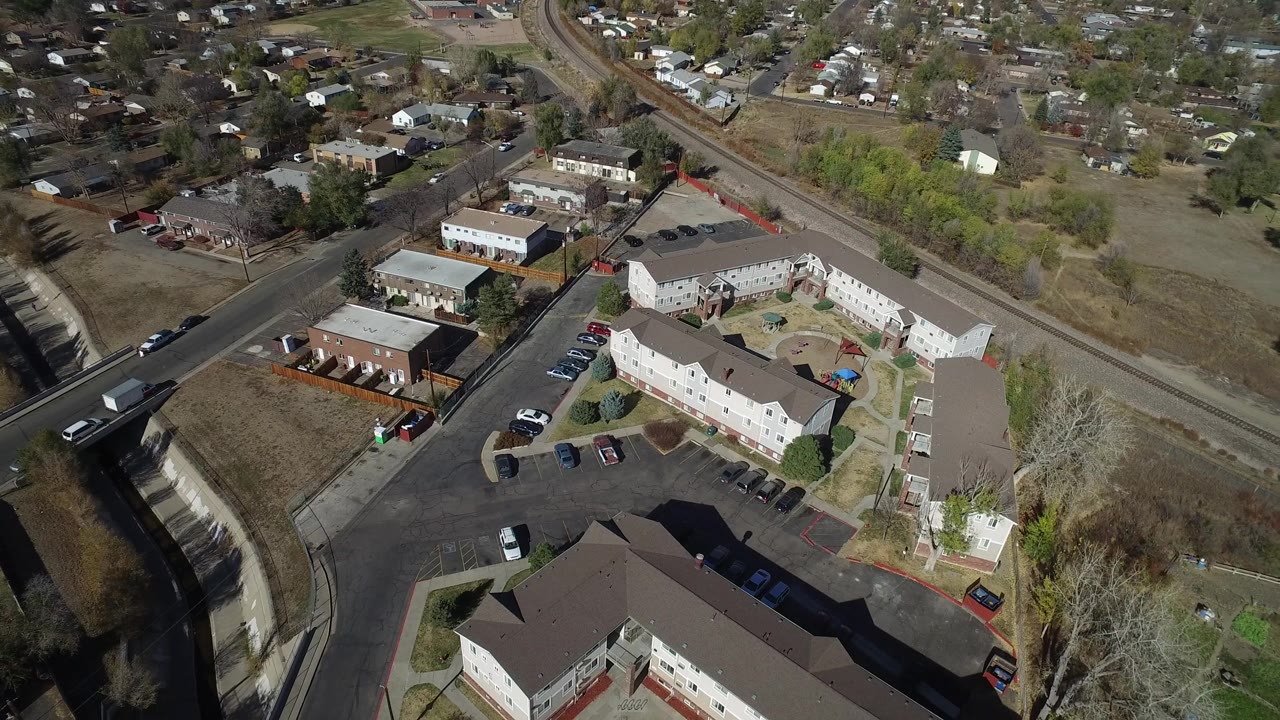 Aerial frame over Westminster, Colorado, with a low-rise apartment courtyard, parking rows, a road along a drainage channel, distant homes, and no visible people.