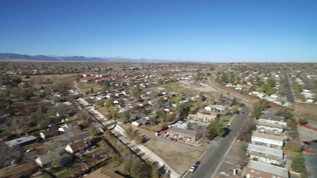 Aerial frame in Westminster, Colorado looking across rooftops, a broad road, small parking areas, tree cover, distant hills, and no visible people.