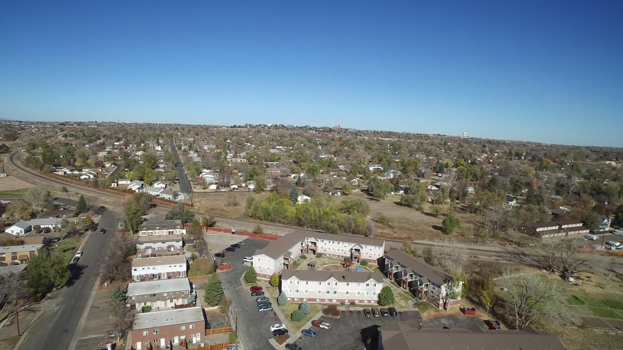 Westminster, Colorado aerial view with apartment buildings in the lower frame, single-family homes beyond, open ground, parked vehicles, and no visible people.